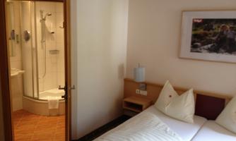 A simple hotel room with two single beds and a bathroom with a shower. The walls are painted in neutral colors, and a picture hangs on the wall.