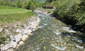 A clear river flows through a green landscape with meadows and trees. Gentle hills can be seen in the background.