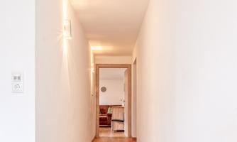 A bright hallway with white walls and a wooden floor. At the end of the hallway, a living room can be seen.