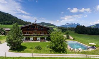 A charming wooden house with a swimming pool in a green landscape. In the background, mountains and a blue sky can be seen.