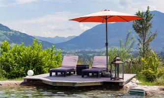A relaxing area by the water with two lounge chairs and a large sun umbrella. In the background, there are mountains and a green landscape.