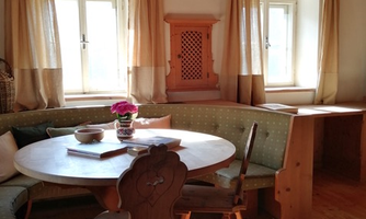 A cozy dining area with a round table and wooden chairs. Large windows let in plenty of daylight, and the walls are adorned with soft curtains.
