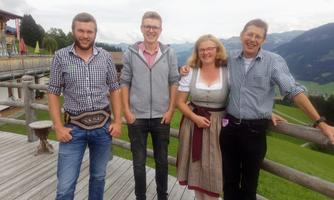 A group of four people stands smiling on a wooden terrace with a beautiful mountain landscape in the background. They are wearing traditional and casual clothing that emphasizes their friendly character.