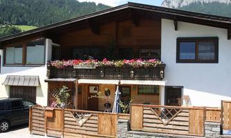 A charming house with a balcony full of flowers. In the foreground, there is a wooden fence, and in the background, green mountains can be seen.