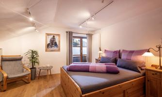 A cozy bedroom with a large wooden bed and pillows. An armchair and a window with a view outside complete the furnishings.