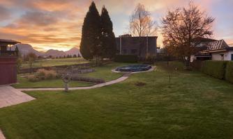 A beautiful garden with green grass, a pond, and trees. In the background, there is a house and the sky glows in warm colors during sunset.