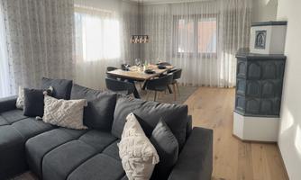 A modern living and dining room with a large, comfortable sofa and an elegant dining table. The large windows let in plenty of daylight and create a bright, inviting atmosphere.