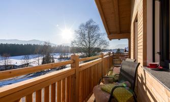A sunny balcony with a wooden railing and two chairs. In the background, a peaceful winter landscape with trees and a blue sky can be seen.