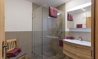 A modern bathroom with a glass shower and a wooden sink. Red towels hang on the wall and a chair stands next to the shower.