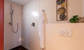 A modern shower with a handheld showerhead and bright, simple wall cladding. A towel and a plant are also visible.