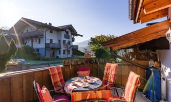 A cozy outdoor area with a round table and colorful chairs. In the background, a sunny house and a green landscape can be seen.