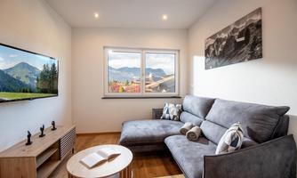 A cozy living room with a gray sofa and a round table. Large windows offer a view of the surrounding mountains.