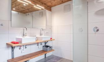 A modern bathroom with two sinks and an open shower. The walls are light-tiled and the wooden ceiling adds a warm atmosphere.