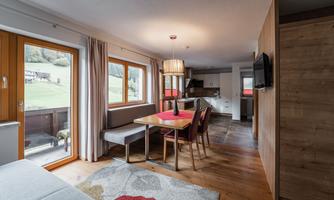 A modern apartment with a dining table and chairs. Large windows offer a view of the green landscape.