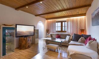 A cozy living room with a wooden floor and wooden ceiling. There is a large sofa, a television, and a window with curtains.