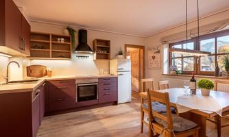 A modern kitchen with wooden furniture and a dining table. Large windows provide bright light and a cozy atmosphere.