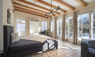 A bright, modern bedroom with a wooden beam ceiling and large windows. The bed is cozy, decorated with a blanket and pillows.