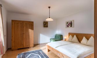 A bright bedroom with a double bed, a wardrobe, and a cozy sitting area. The floor is made of wood, and a carpet adds a warm atmosphere.