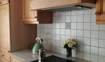 A modern kitchen with wooden cabinets and bright tiles. On the countertop, there is a kettle and a vase with flowers.