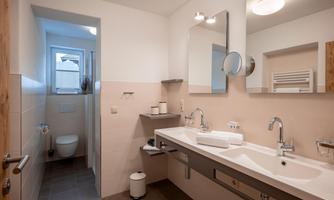 Modern bathroom with a large mirror, two sinks, shower, toilet, and window.