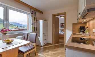 A modern kitchen with wooden flooring and bright interior. On the table, there is a fruit bowl and a vase of flowers, while the window offers a beautiful view of the mountains.