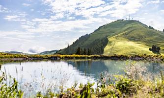 A quiet mountain lake with green meadows and gentle hills in the background. The sky is blue and lightly clouded.