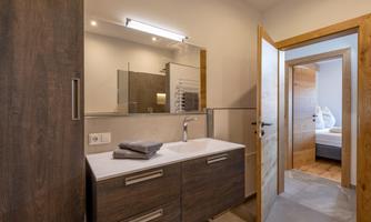 A modern bathroom with wooden cabinets and a bright mirror. In the background, another room can be seen.