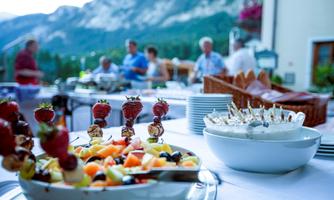 A festively laid table with colorful fruit skewers and a bowl of snacks. In the background, people and a beautiful mountain landscape can be seen.