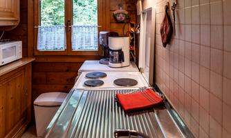 A cozy kitchen with wooden paneling and large windows. On the countertop are a stove, a coffee machine, and a dish towel.