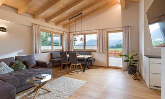 A modern living room with a wooden ceiling and large windows. There is a cozy sofa area and a dining table with chairs.