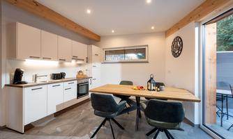 A modern kitchen with a large table and stylish chairs. The light emphasizes the bright walls and the wooden details.