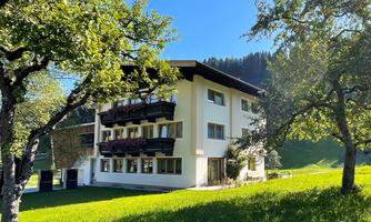 A modern house surrounded by green meadows and trees. The clear sky and the mountains in the background give the surroundings an idyllic atmosphere.