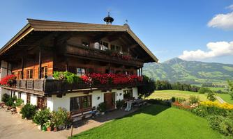 A traditional wooden house with a sheltered balcony and blooming flower boxes. In the background, there are green hills and a clear sky.