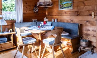 A cozy wooden cabin with a round table and a bench. Sunlight falls on the furnishings, and there are decorative elements on the table.