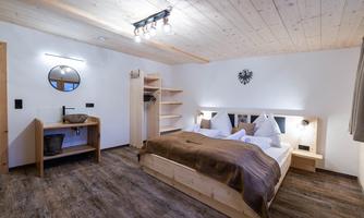 A modern room with a double bed, wooden floor, and minimalist decor. The space is bright and cozy, featuring a large mirror and an open wardrobe.