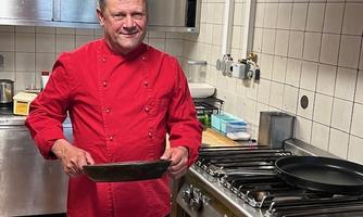 A chef in a kitchen is wearing a red chef's jacket and holding a pan in hand. In the background, cooking equipment and utensils are visible.