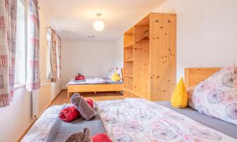 A bright, inviting bedroom with two beds and a large window. There is a wooden shelf with colorful towels on the beds.