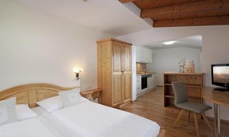 A bright room with two single beds and wooden furniture. There is a small table and a television, as well as a kitchenette in the background.