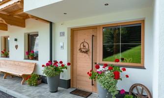 An inviting entrance to the house with a wooden front door and large planters full of flowers. Next to the door stands a cozy wooden bench.