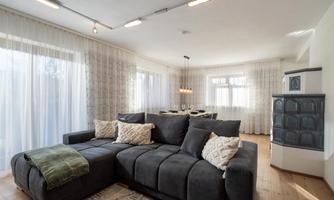 A modern living room with a large, comfortable sofa and bright curtains. The room is bright and inviting, with a dining table in the background.