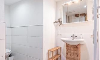 A modern bathroom with a white wall and wooden shelves. A sink with a mirror and a shower is also visible.