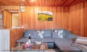 A cozy living room corner with a gray couch and a glass table. Wooden paneling hangs on the wall along with a picture of a landscape.