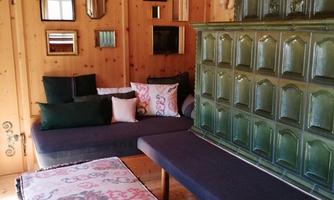 A cozy living space with wooden paneling. Cushions lie on the sofa, and a green stove stands as a highlight against the wall.
