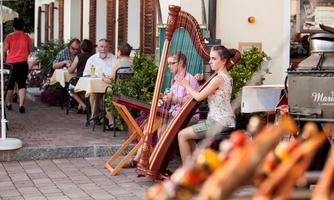 A young woman plays the harp in a busy café. Guests are sitting at tables and enjoying the atmosphere outdoors.