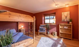 A cozy bedroom with a wooden loft bed and rustic furnishings. The walls are painted in a warm red tone and there is a small reading area with a plant.
