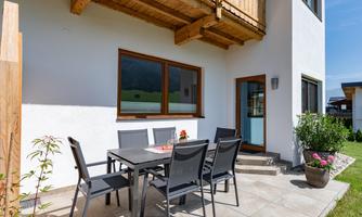 A sunny terrace with a large table and modern chairs. The view leads to a cozy residential house and a green environment.