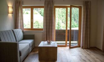 A bright living room with a gray sofa and a wooden table. Large windows with curtains provide a view of the greenery.