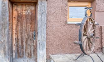 An old wooden door with the inscription "21 KH 25" and a historical wagon wheel next to it. The wall is painted in a dark brown tone, and a small window is visible.