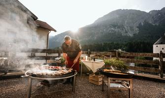 A grill master prepares various dishes outdoors, surrounded by a picturesque mountain landscape. The smoke rises into the air as the sun shines in the background.