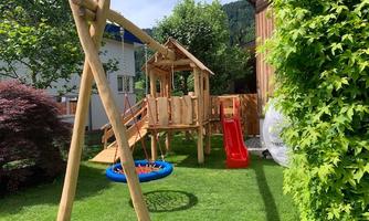 A beautiful playground with a wooden swing, a slide, and a playhouse. The area is surrounded by green grass and bushes.